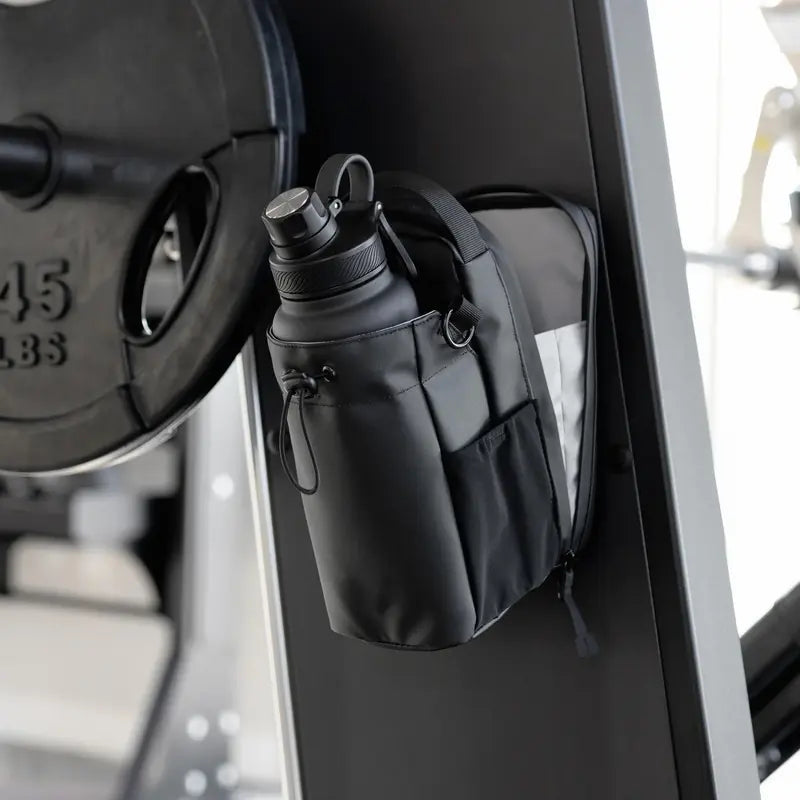 Un sac magnétique Wizzi Tn noir avec une bouteille d'eau dans sa poche latérale est suspendu à un appareil de musculation, avec une plaque de 45 livres à proximité dans le centre de remise en forme.